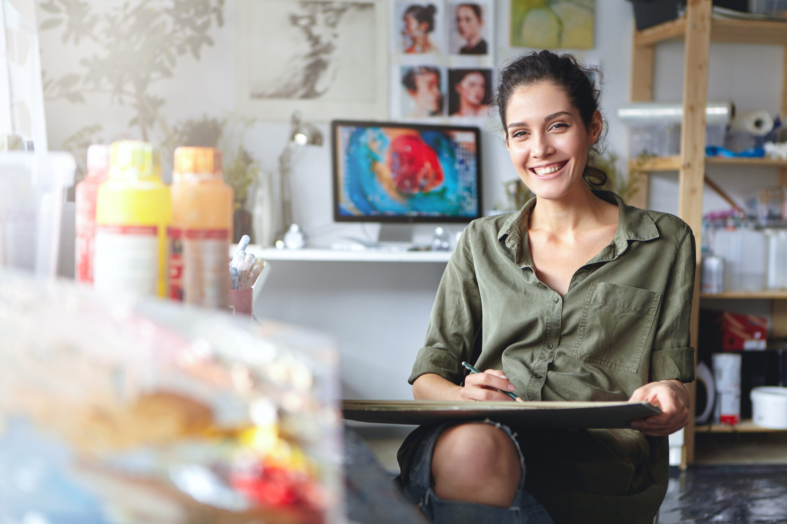 Smiling female artist wearing casual clothes sitting in her cabinet with sketches and colorful paint, having happy expression while being glad to create beautiful picture. Painter working in workshop